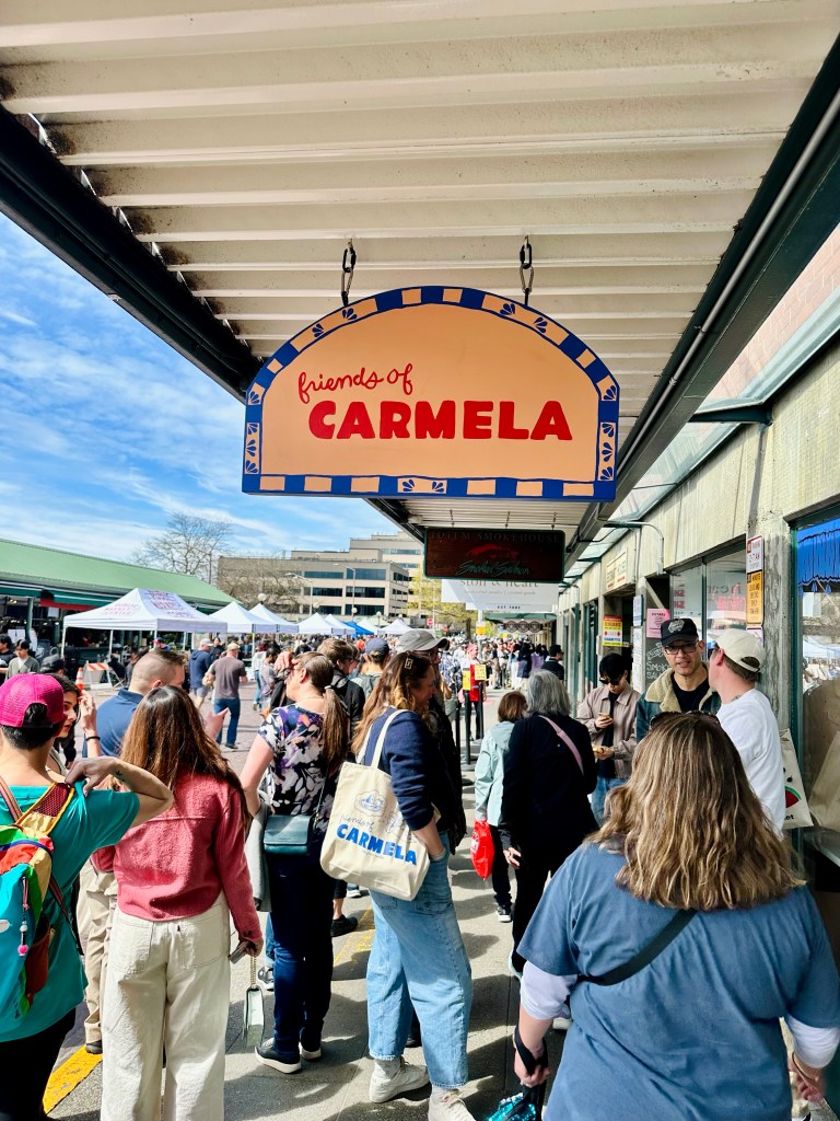 A bustling outdoor market scene under a sign that reads 'friends of CARMELA', with diverse crowds of people shopping and walking among various stalls.