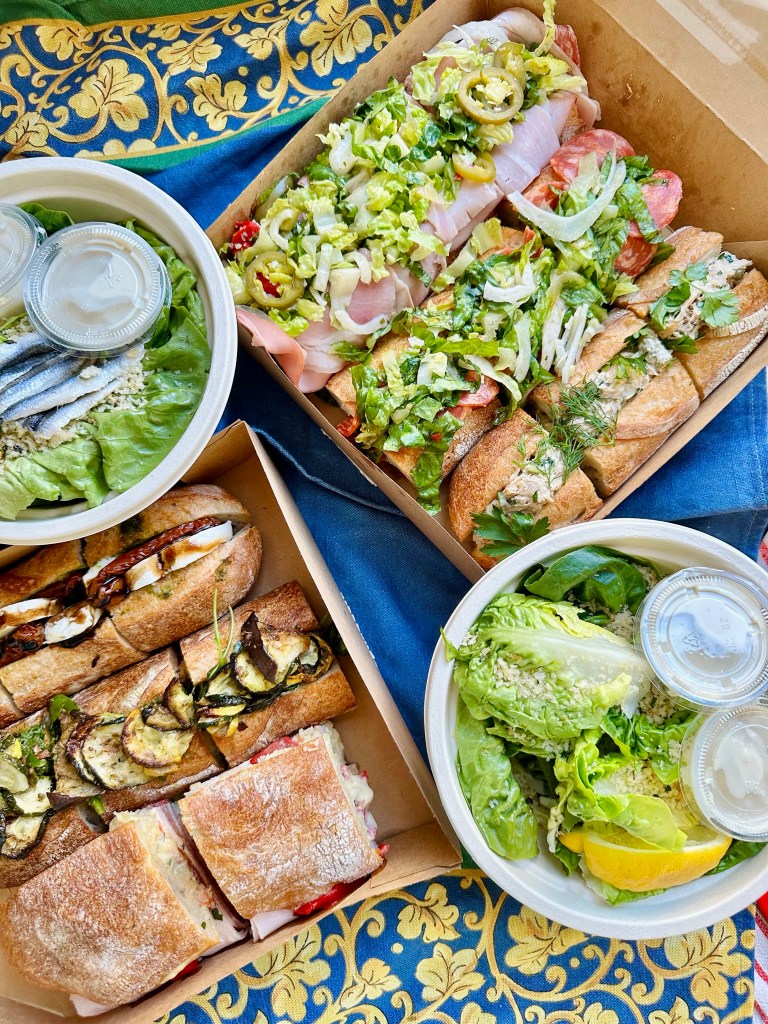An assortment of sandwiches with various toppings including meats, vegetables, and sauces, placed in cardboard boxes, alongside a bowl of mixed greens with dressing on a decorative tablecloth.