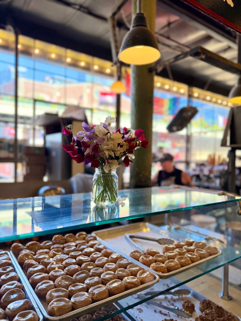 A glass display case filled with sweet pastries, with a vase of colorful flowers in the foreground and a bustling market atmosphere in the background.