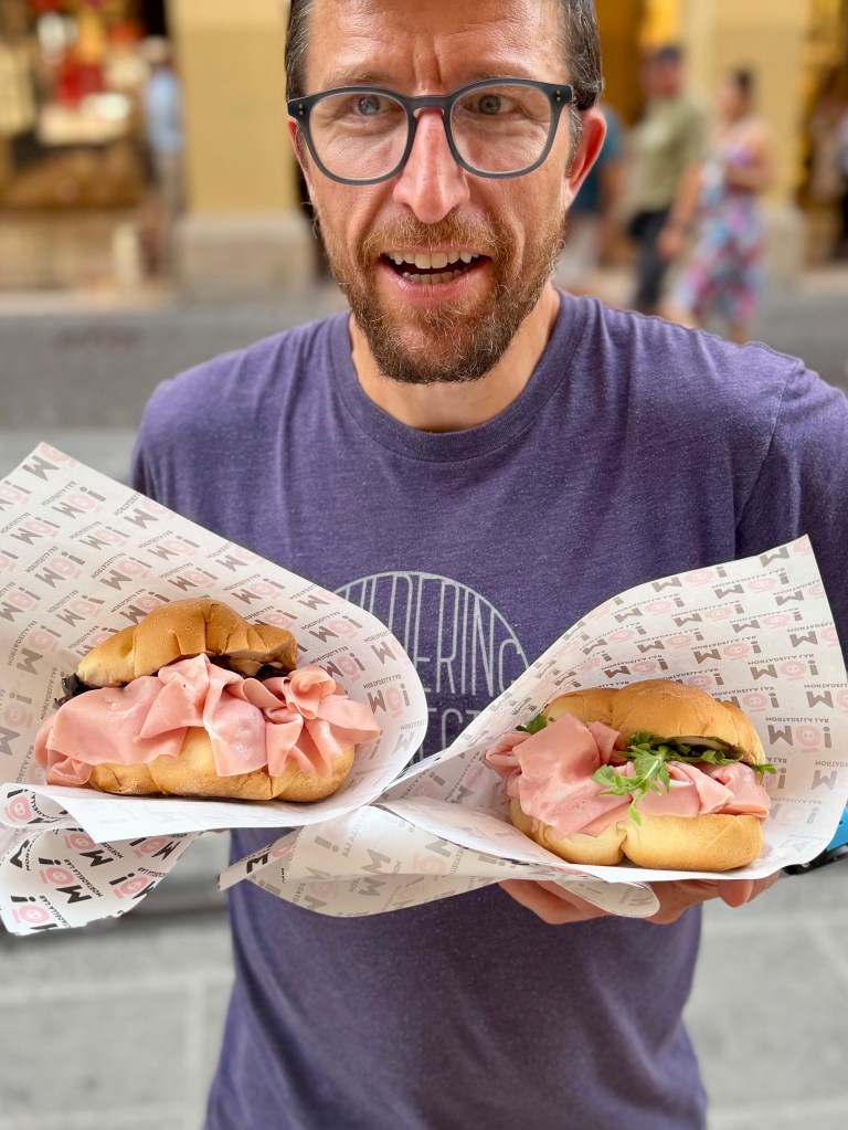 A man holding two sandwiches wrapped in paper, filled with layers of ham and some greenery, standing outdoors in a bustling street.