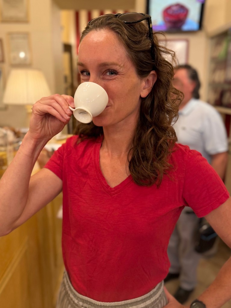 Woman in a red shirt holding a small espresso cup and smiling.