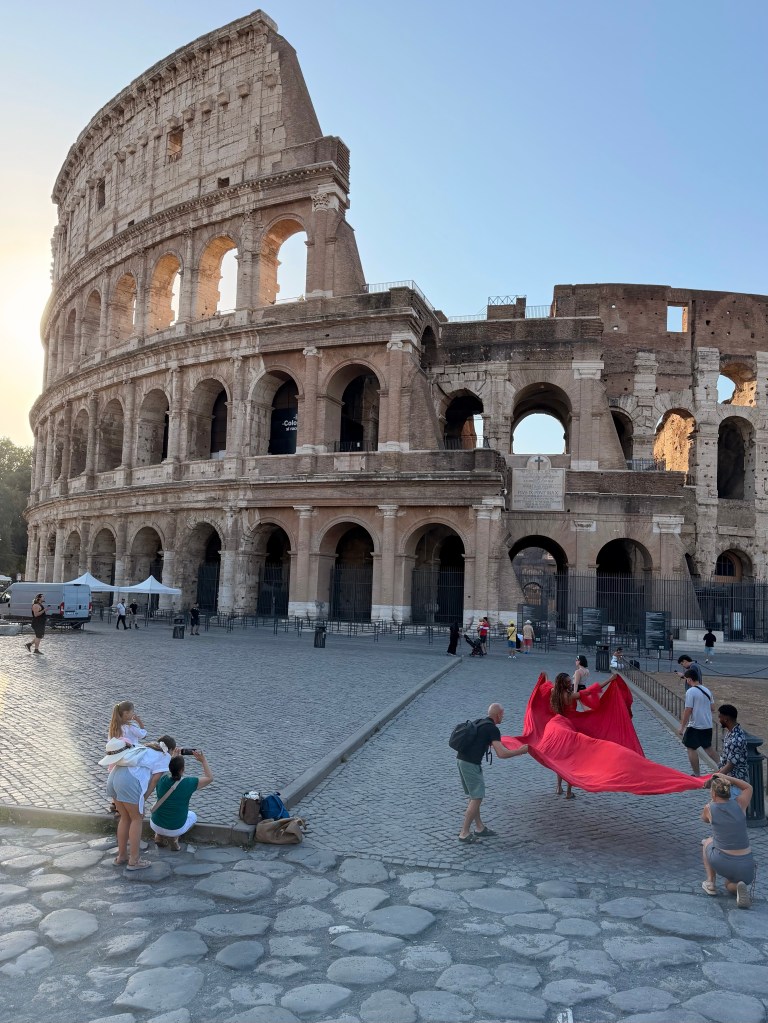 A large group of people near the Colosseum in Rome, some posing for photos while others hold a red fabric, on a cobblestone square with a clear sky.