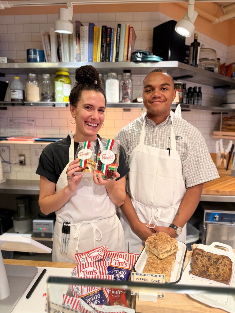 A smiling woman and man stand behind a counter in a cafe, holding boxes of Italian cookies. The woman wears a black shirt and apron, while the man is in a plaid shirt and white apron. They are surrounded by baked goods and snacks like potato chips and coffee cake.