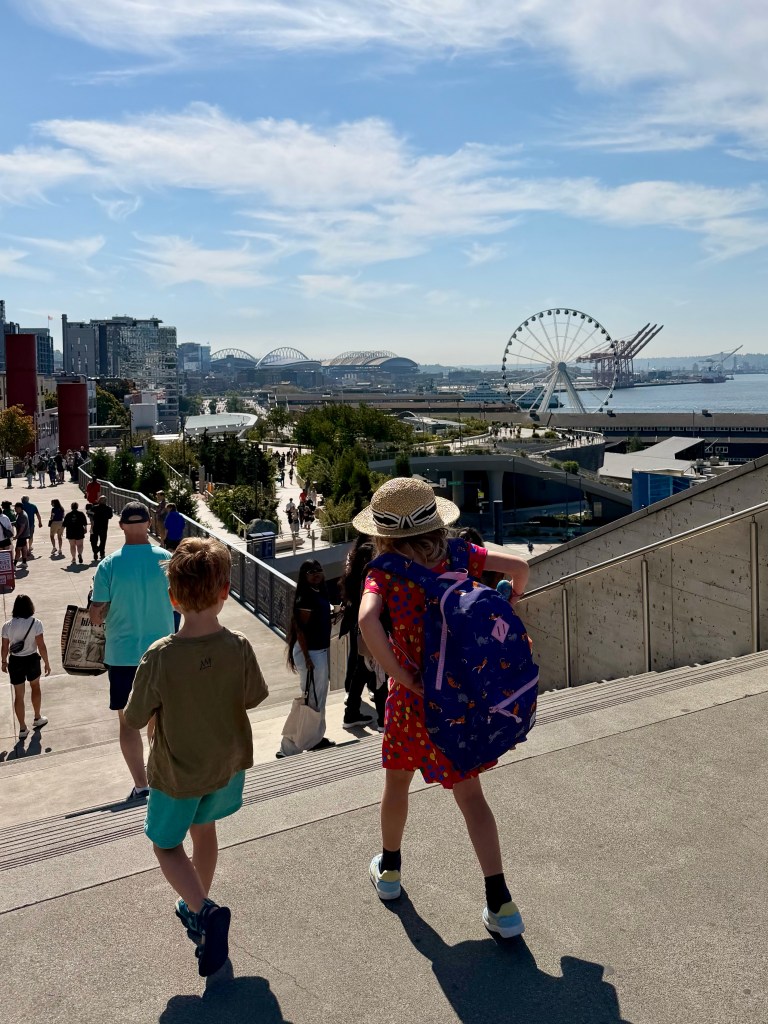 Two children walking down a staircase in an outdoor setting, with a waterfront view featuring a ferris wheel and buildings in the background.