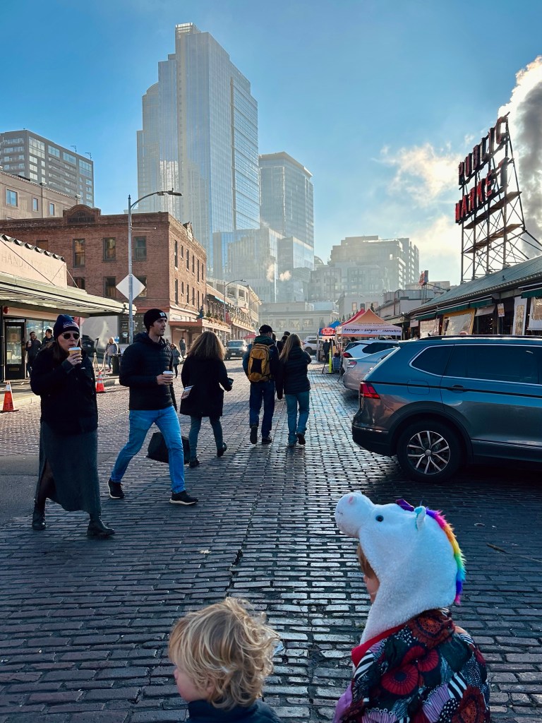 A busy street scene at a marketplace with people walking, modern buildings in the background, and two children in the foreground; one wearing a unicorn hat.