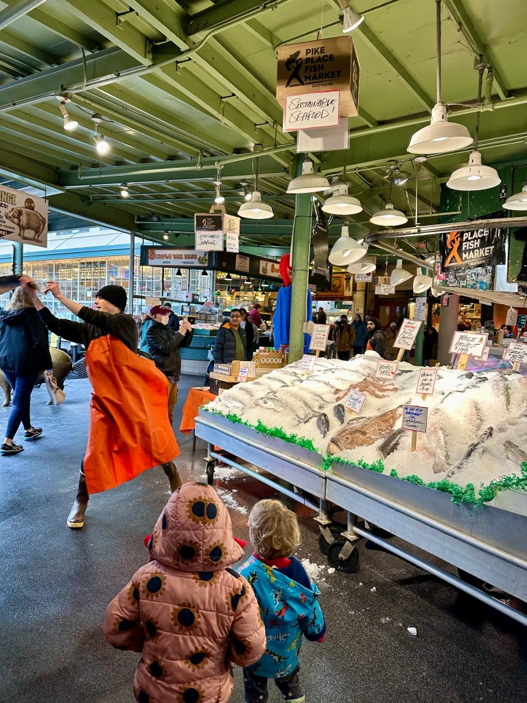 A lively scene at Pike Place Fish Market showcasing a vendor throwing a fish, with children watching excitedly. The market features a display of ice-covered seafood and various signage promoting sustainable seafood.