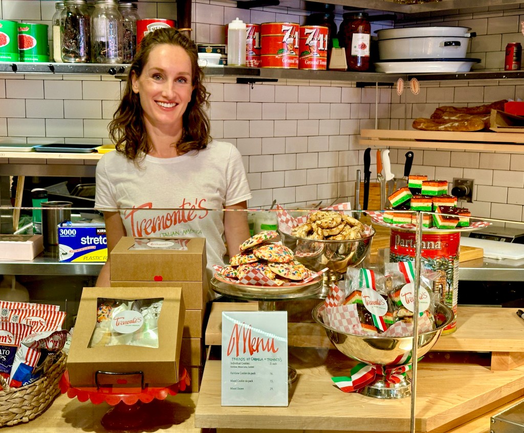 A smiling woman stands behind a counter displaying various baked goods, including cookies and desserts, at Tremonte's Italian bakery. The display features colorful treats, packaging with the Tremonte's logo, and a menu on the counter.