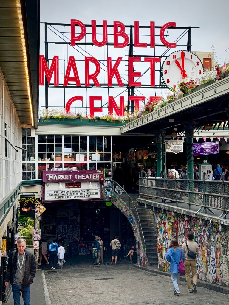 View of the iconic Public Market Center sign with a clock, located at Pike Place Market in Seattle, featuring pedestrians and market stalls in the foreground.