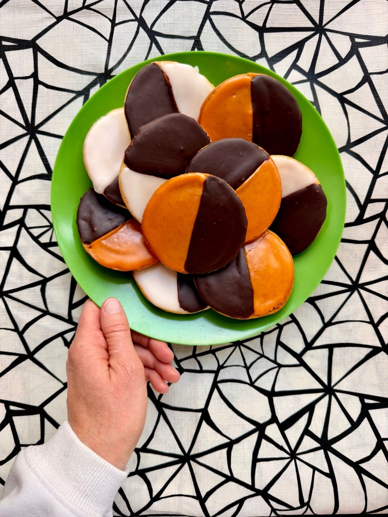 A hand holding a green plate filled with round cookies in various colors, including orange, black, and white, placed on a spiderweb-patterned tablecloth.