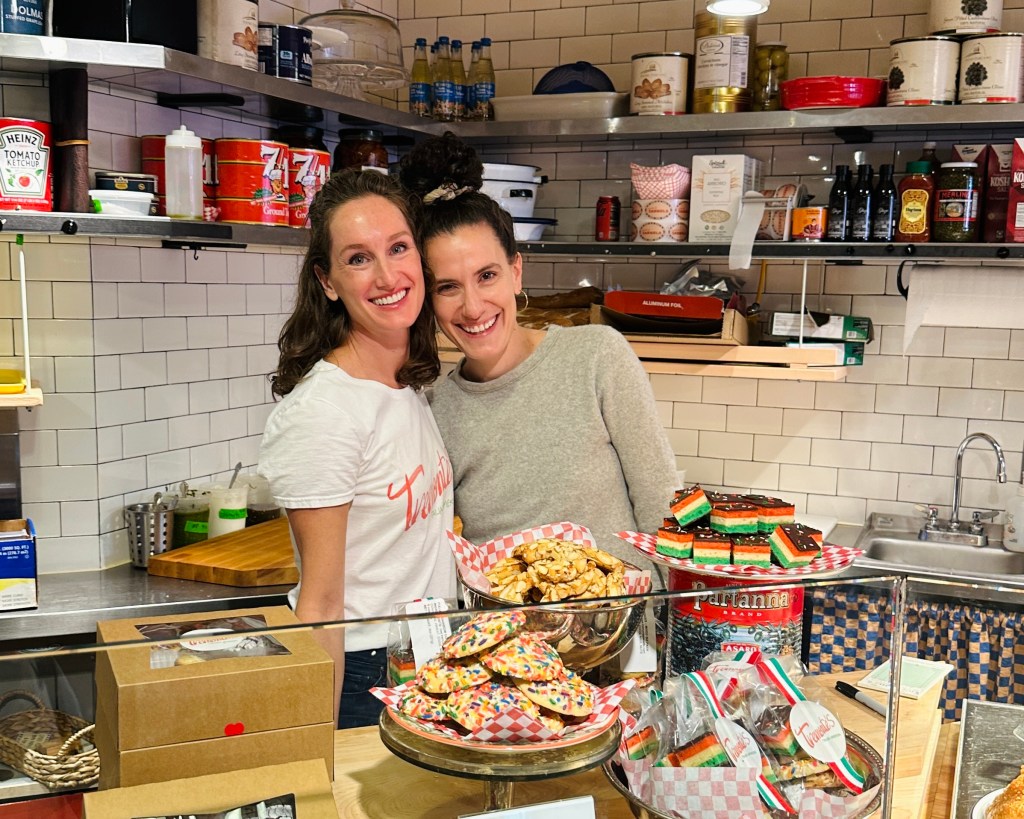 Two women smiling behind a dessert counter filled with various baked goods, including colorful cookies and Italian pastries, in a cozy café setting.